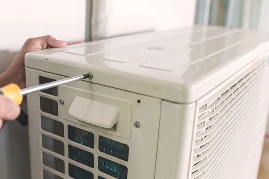 Hand holding a screwdriver and removing a screw on the side of an outdoor air conditioning unit.
