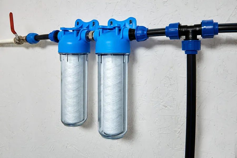 Dual water filtration unit with blue housings and clear cartridge canisters mounted to a wall, connected to piping with a red shut-off valve on the left.