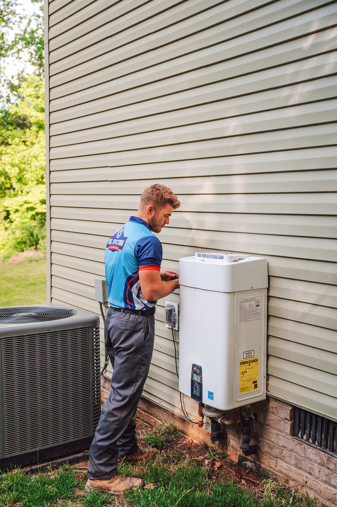 Plumber in uniform working on an outdoor tankless water heater next to an air conditioning unit in a residential area with siding and grassy surroundings.