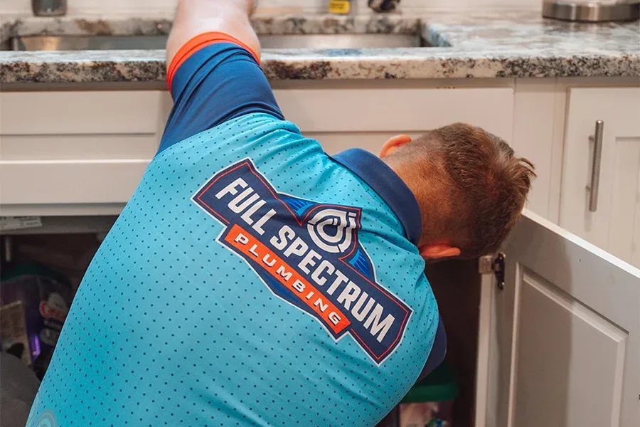 Plumber in a turquoise Full Spectrum Plumbing shirt kneels under a kitchen sink, inspecting pipes under the counter.