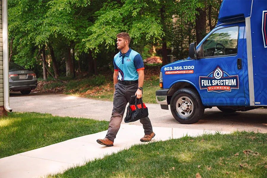 Plumber in a blue polo walks on a sidewalk, carrying a tool bag beside a blue Full Spectrum Plumbing van parked on a residential street.