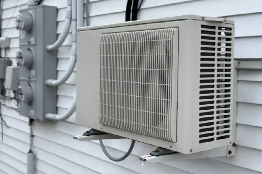 Outdoor air conditioner condenser unit mounted on a white siding wall with electrical panels nearby