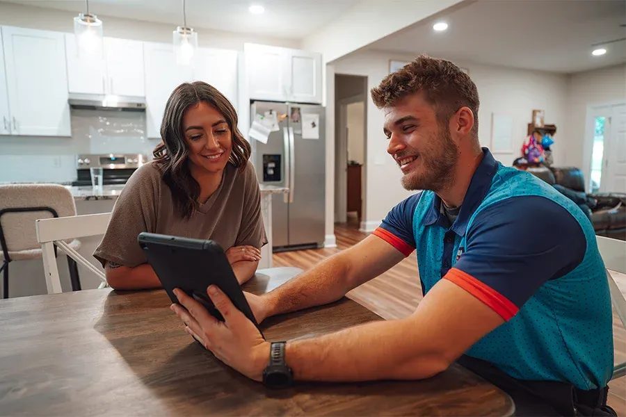 Two adults sit at a dining table in a bright kitchen, smiling while looking at a tablet together on the table.