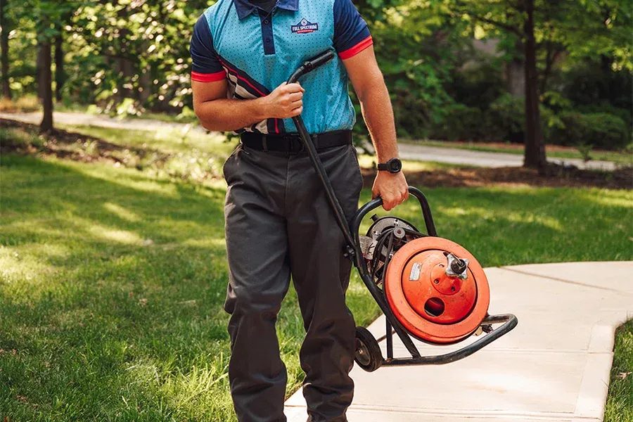 Person in a teal polo shirt pulls an orange extension cord reel on a wheeled stand along a park sidewalk.