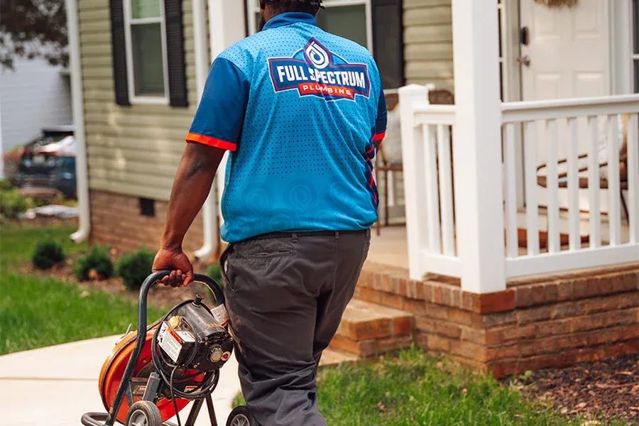Plumber in a blue Full Spectrum Plumbing shirt pushes a hose reel along a residential sidewalk toward a house on a front yard job site (informative)