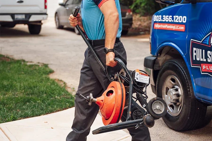 Plumber in a teal shirt walks on a driveway carrying a portable drain-cleaning machine beside a blue service van with company branding.