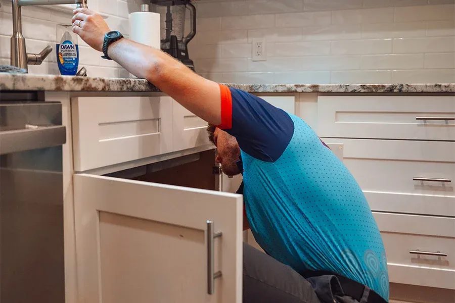 Man kneeling under a kitchen sink, hand reaching to faucet area for maintenance or repair.