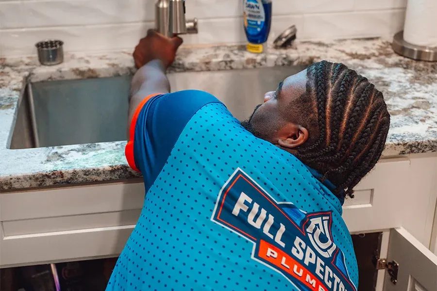 Plumber leaning over a kitchen sink, turning the faucet with a blue logo shirt reading 'Full Speed Plumbing' visible on the back.