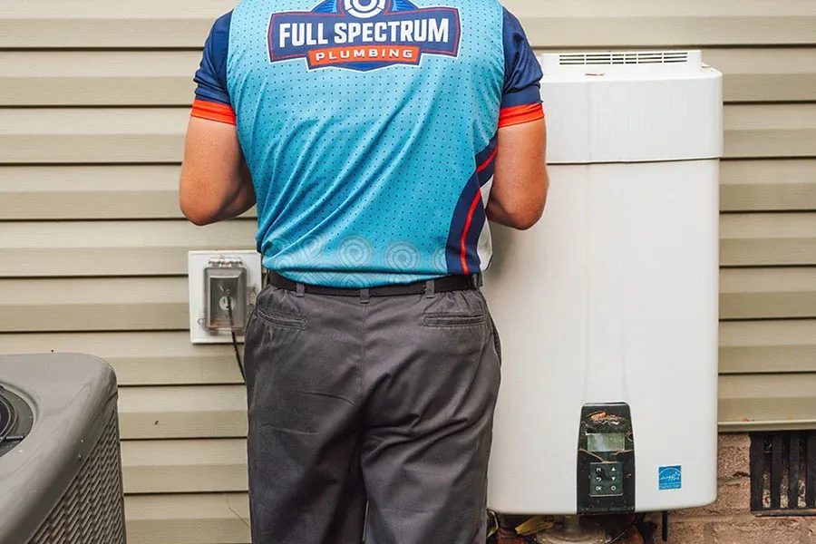 Plumber in a blue Full Spectrum Plumbing jersey seen from behind, standing beside an outdoor water heater on a beige house exterior.