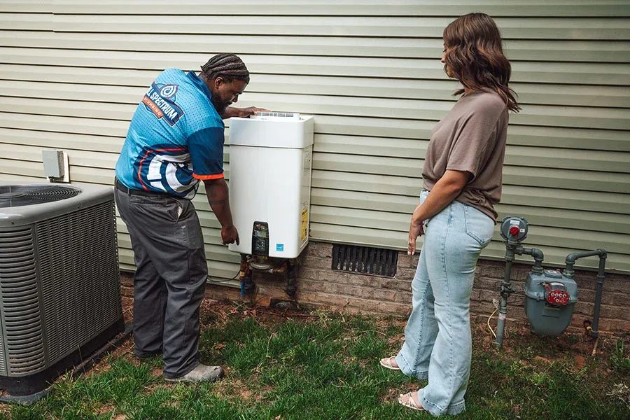 Technician in a blue jersey installs or inspects an outdoor wall-mounted water heater as a woman watches nearby on a grassy yard beside the house exterior.